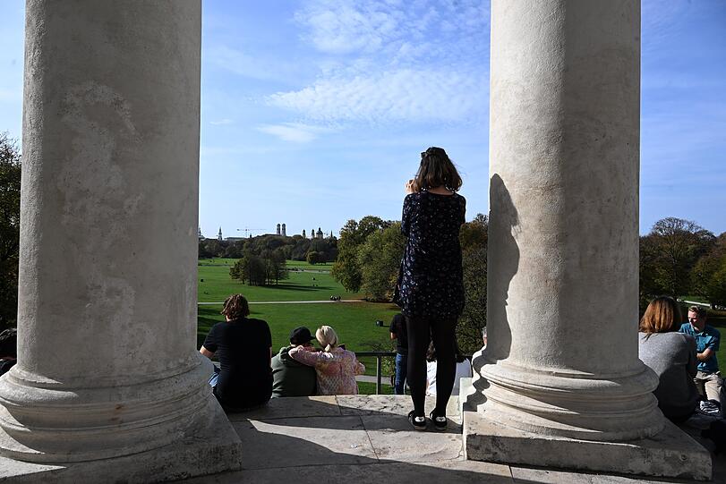Eine Frau schaut bei schönem Wetter vom Monopteros aus auf die Silhouette der Landeshauptstadt München.