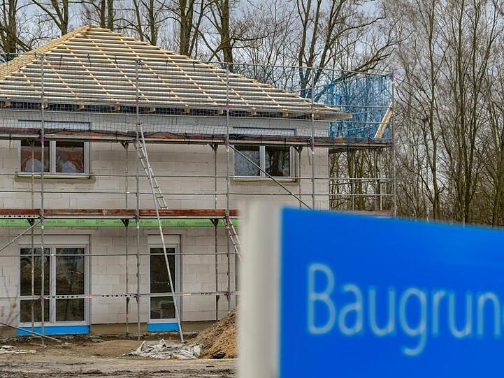 Ein Schild mit der Aufschrift "Baugrundstücke" steht am Rande eines Baufeldes mit einem Rohbau eines Eigenheimes.