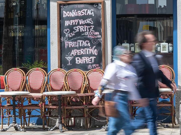 Passanten gehen an zusammengestellten Tischen und Stühlen eines geschlossenen Cafes vorbei. Foto: Peter Kneffel/dpa/Archivbild