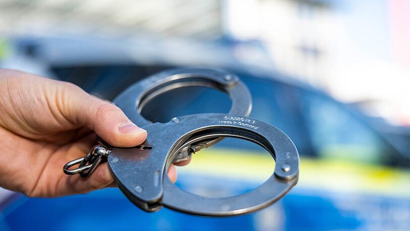 Eine Hand hält Handschellen vor einen Streifenwagen der Polizei.