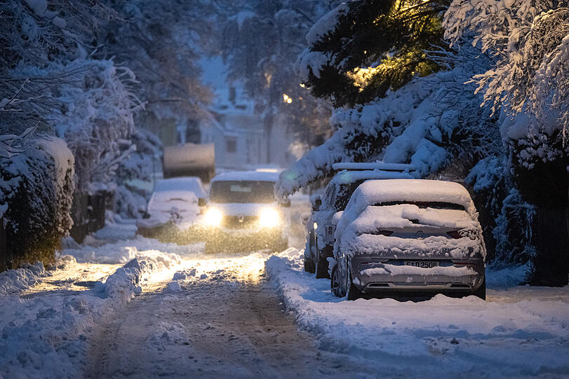 Ein Transporter f&auml;hrt mit Licht durch die verschneite Stra&szlig;e, w&auml;hrend parkende Autos unter einer dicken Schneeschicht liegen.