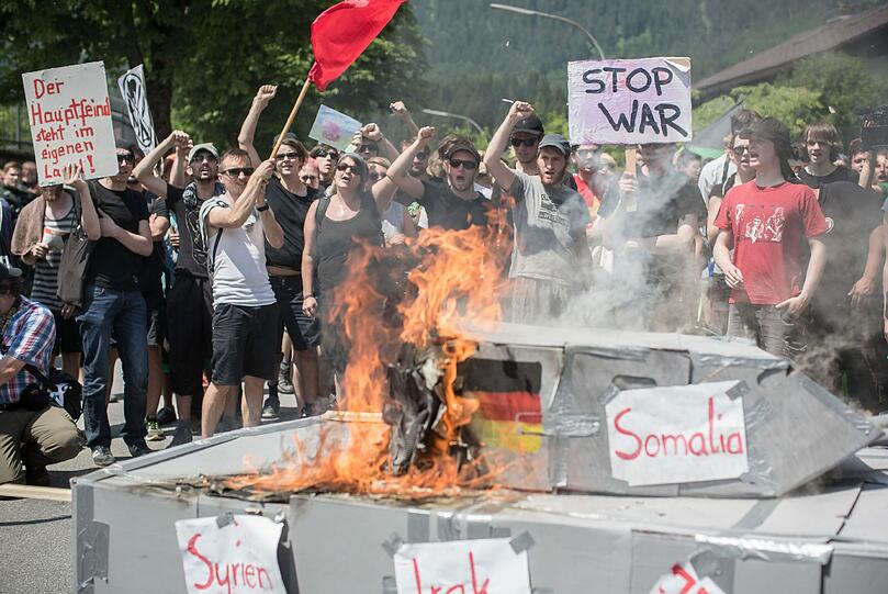 Fotografen, Reporter und Kameraleute umringen einen brennenden Panzer aus Pappe, auf dem die Ländernamen der Krisenherde Lybien, Mali, Kurdistan und Ukraine stehen, während einer Demonstration vor dem Marshall Center for European Security Studies in Garmisch-Partenkirchen.