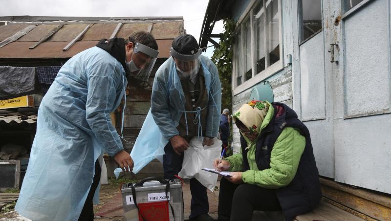 Zwei Wahlhelfer stehen neben einer Frau, die auf den Stufen vor einem Haus sitzt und Dokumente ausfüllt.