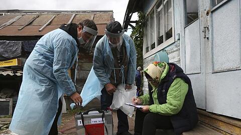 Zwei Wahlhelfer stehen neben einer Frau, die auf den Stufen vor einem Haus sitzt und Dokumente ausfüllt.