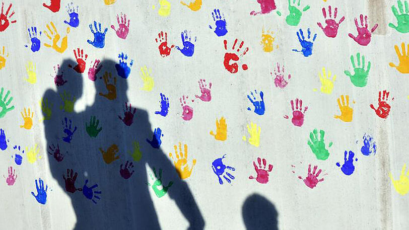 Ein Mann mit einem Kind auf dem Arm und einem an der Hand wirft einen Schatten auf eine mit bunten Handabdrücken bemalte Wand einer Kindertagesstätte.