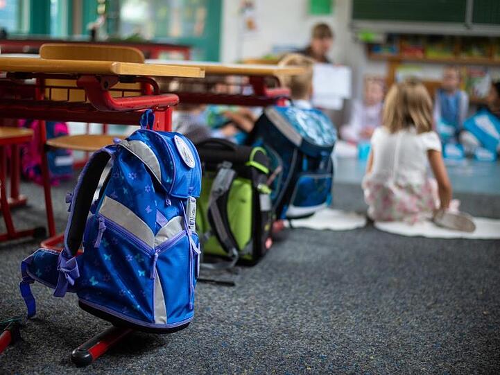 Schulranzen hängen und stehen in einem Klassenzimmer. Im Hintergrund sitzen Schulkinder am Boden. Foto: Philipp Schulze/Archiv