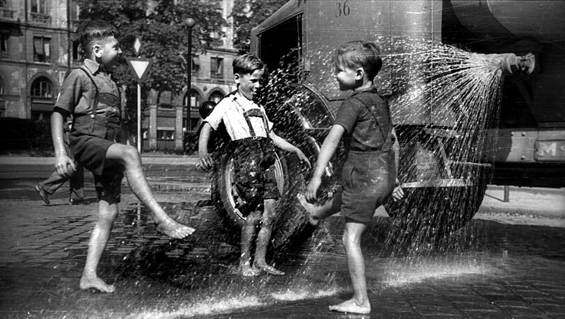 Hochsommer in M&uuml;nchen 1956: Ein Spritzenwagen der Stadtreinigung bringt f&uuml;r diese drei Buben in kurzer Lederhosn eine &auml;u&szlig;erst willkommene Abk&uuml;hlung.