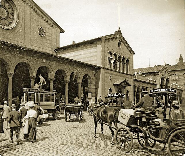 Das ist der alte Münchner Hauptbahnhof von Friedrich Bürklein im Jahr 1910. Vor ihm stehen Pferdekutschen und Omnibusse der Luxushotels (hier: Bayerischer Hof, Deutscher Hof, Roter Hahn). Während des Zweiten Weltkriegs wird er stark beschädigt, 1949 werden die einsturzgefährdeten Gebäude gesprengt.