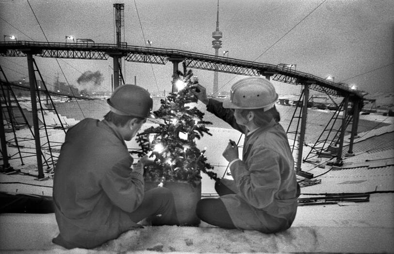 Dezember 1970: Bauarbeiter entzünden einen kleinen Christbaum auf der Haupttribüne des Olympiastadions, das noch eine Großbaustelle ist.