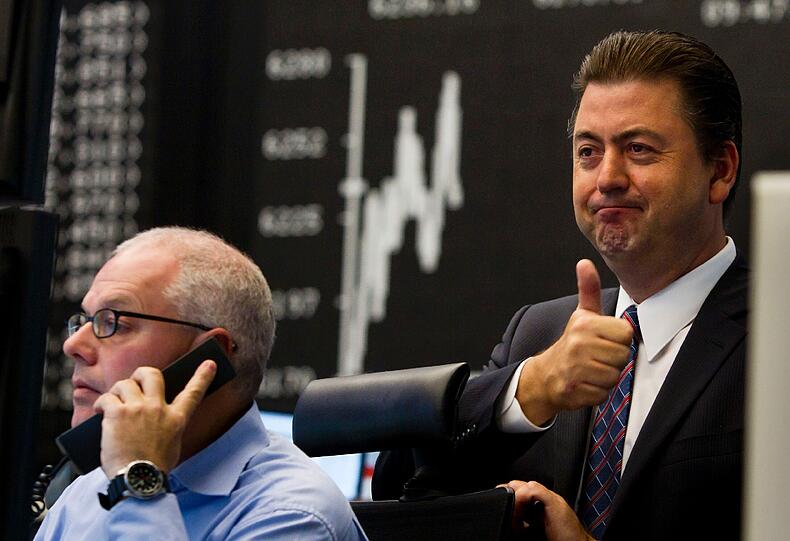 Robert Halver, Kapitalmarktexperte der Baader Bank, zeigt in der Börse in Frankfurt am Main vor der Anzeigetafel für den DAX-Index einem Kollegen den erhobenen Daumen. Es geht bergauf.