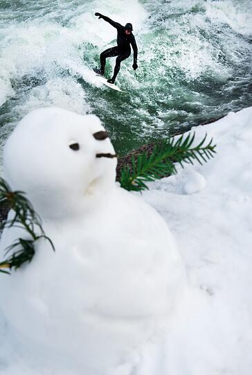 Abseits der Schneeballschlacht reitet ein Surfer die Eisbachwelle. Stiller Beobachter: Der Schneemann.