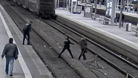 Ein Bild der Überwachungskamera zeigt die Situation. Rechts die Frau, die übers Gleis läuft, hinter ihr die Polizisten. Im Hintergrund der einfahrende Zug. Ein Bild der Überwachungskamera zeigt die Situation. Rechts die Frau, die übers Gleis läuft, hinter ihr die Polizisten. Im Hintergrund der einfahrende Zug.