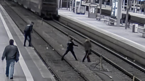 Ein Bild der Überwachungskamera zeigt die Situation. Rechts die Frau, die übers Gleis läuft, hinter ihr die Polizisten. Im Hintergrund der einfahrende Zug.