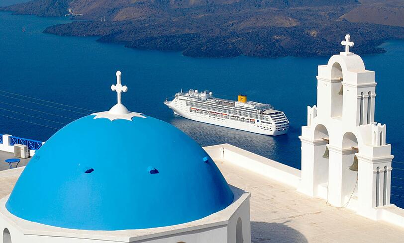 Santorin: Blick über eine der berühmten blauen Kuppeln von Oia, die eines der Wahrzeichen Griechenlands sind, auf ein Kreuzfahrtschiff in der Ägäis.