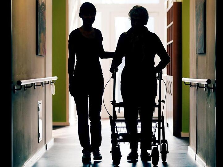 Eine Pflegekraft (l) begleitet die Bewohnerin eines Altenheims beim Gang über den Flur. Laut BA sind im Jahresdurchschnitt 2018 in der Altenpflege auf knapp 3100 arbeitslose Fachkräfte rund 15.300 freie Stellen entfallen.