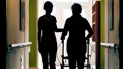 Eine Pflegekraft (l) begleitet die Bewohnerin eines Altenheims beim Gang über den Flur. Laut BA sind im Jahresdurchschnitt 2018 in der Altenpflege auf knapp 3100 arbeitslose Fachkräfte rund 15.300 freie Stellen entfallen.