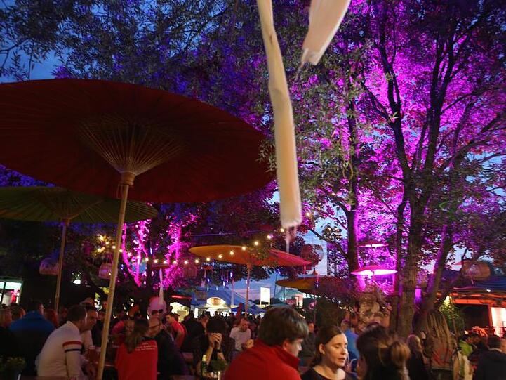 Menschen stehen bei dem Tollwood Festival in München an einer beleuchteten Bar unter freiem Himmel. Foto: Stephan Jansen/dpa/Archivbild