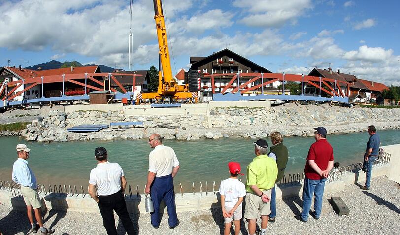 ARCHIV - Passanten beobachten am 05.09.2006 in der oberbayerischen Ortschaft Eschenlohe im Landkreis Garmisch-Partenkirchen am Ufer des Fluss Loisach den Bau von Hochwasserschutzdeichen und einer Brücke.