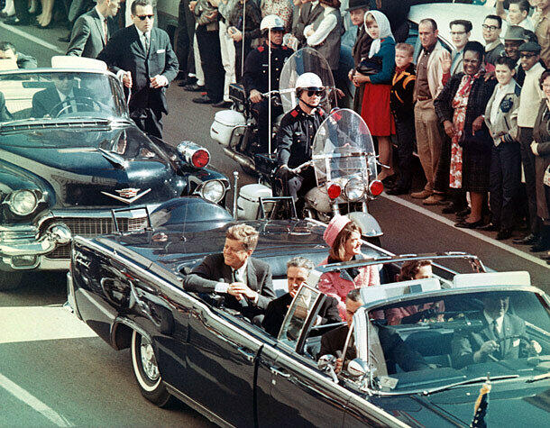 Präsident John F. Kennedy und Gattin Jackie Kennedy lächeln den Menschen am Straßenrand zu. Nur wenige Minuten später, als die Autokolonne die Dealey Plaza erreicht, fallen die tödlichen Schüsse.