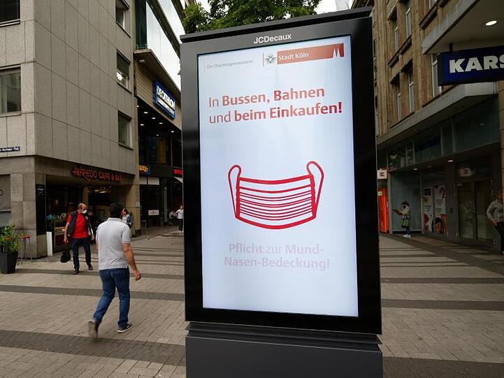 Ein Werbebildschirm weist in der Kölner Innenstadt auf die Maskenpflicht in Geschäften und im Nahverkehr hin.