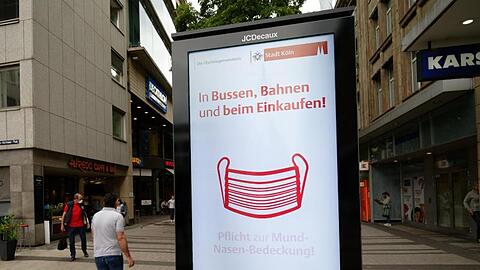 Ein Werbebildschirm weist in der Kölner Innenstadt auf die Maskenpflicht in Geschäften und im Nahverkehr hin.