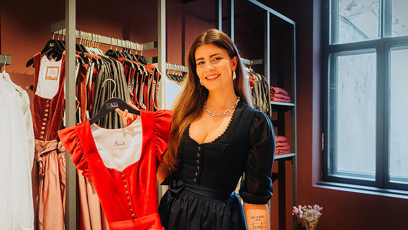 Angelina Kees, selbst in einem eleganten Dirndl in Nachtblau, zeigt ein Stück aus ihrer neuesten Kollektion. Ein rotes Dirndl, das ohne Bluse getragen werden kann.