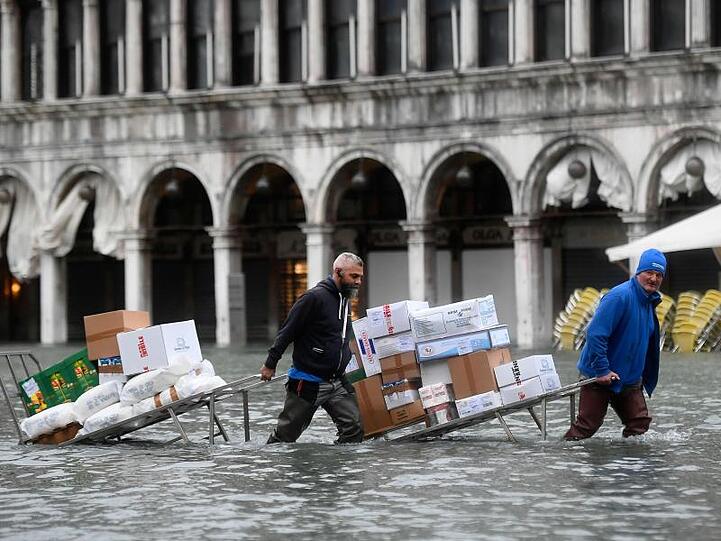 Männer waten in Venedig mit Kartons und Lebensmitteln durch das Hochwasser.