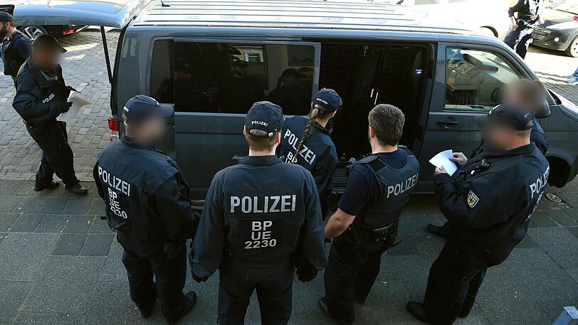 Polizeibeamte und Beamtinnen stehen an einem Einsatzwagen in dem festgenommene Männer sitzen.