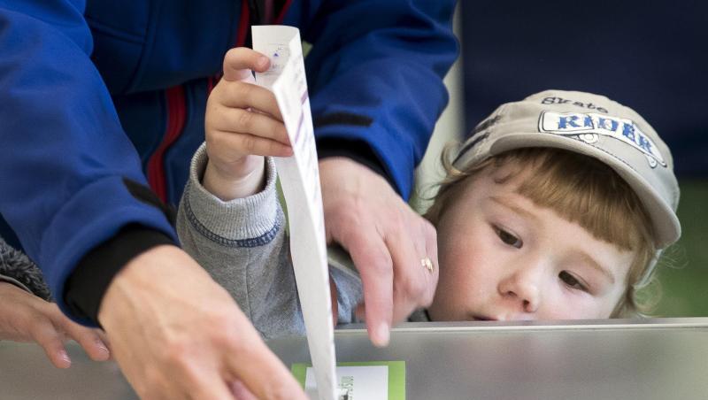 Eine Frau lässt ein Kind ihren Stimmzettel in die Wahlurne stecken.