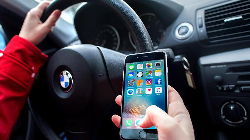 Eine junge Frau sitzt am Steuer und bedient dabei ihr Smartphone.
