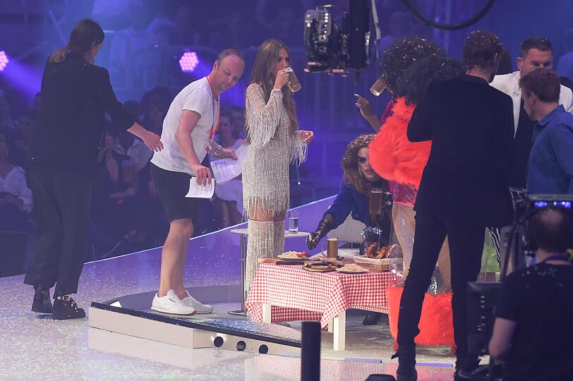 Heidi Klum trink Bier und die Drag Queens essen Würstchen, während ein Mitarbeiter die alten Moderationskarten wegbringt.