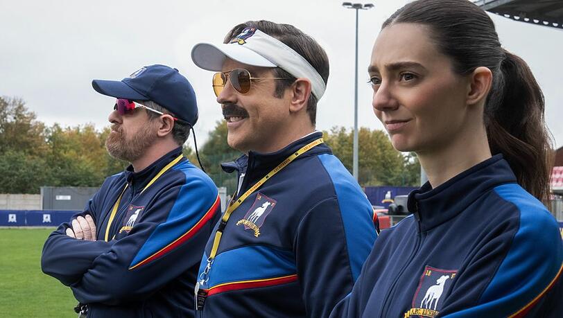 Ted Lasso (Jason Sudeikis, Mitte) sieht sich in Staffel vier einer gänzlich neuen Aufgabe gegenüber, die er mit gewohnter Zuversicht angeht. Ted Lasso (Jason Sudeikis, Mitte) sieht sich in Staffel vier einer gänzlich neuen Aufgabe gegenüber, die er mit gewohnter Zuversicht angeht.